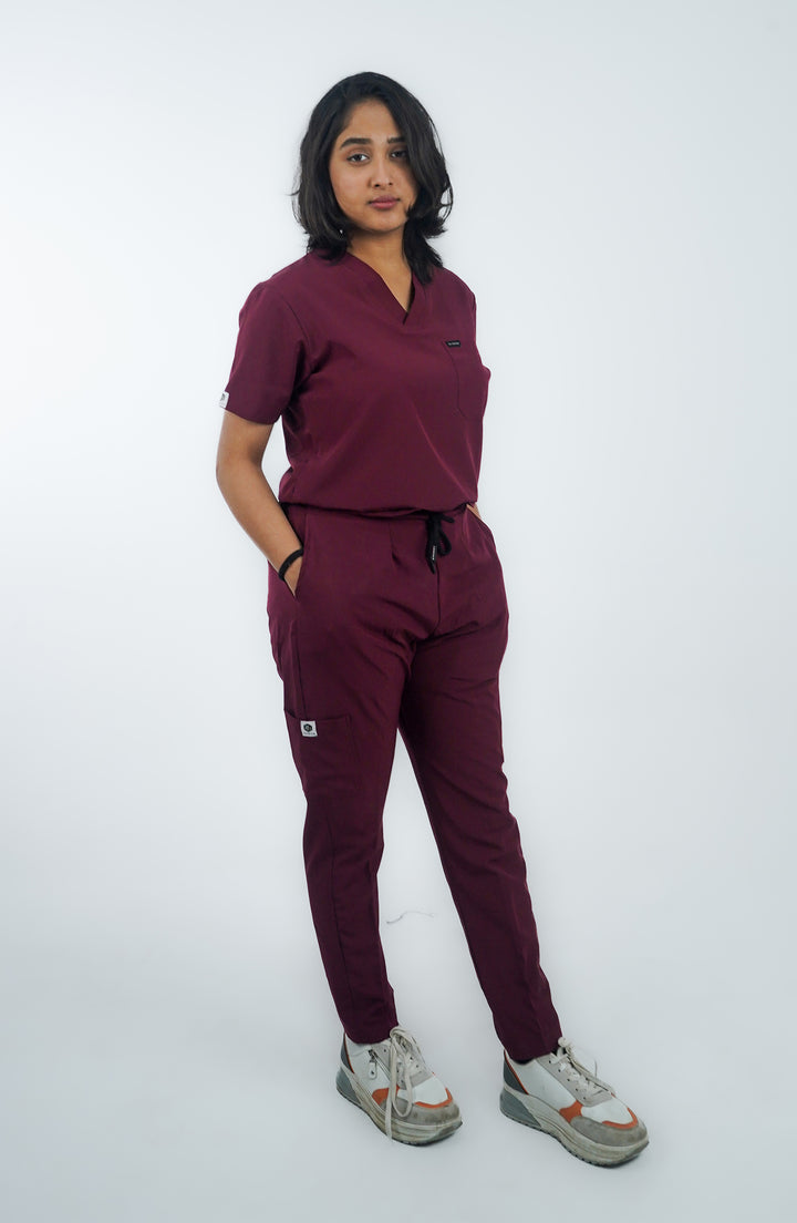 A person standing against a white background, wearing maroon colored scrubs with a V-neck top and button details, paired with white sneakers.