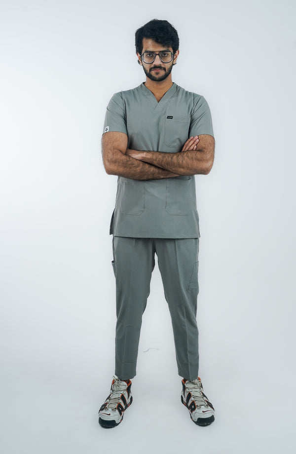 A person standing against a white background, wearing a gray micro-stretch scrub suit with short sleeves and a V-neckline, paired with white sneakers.