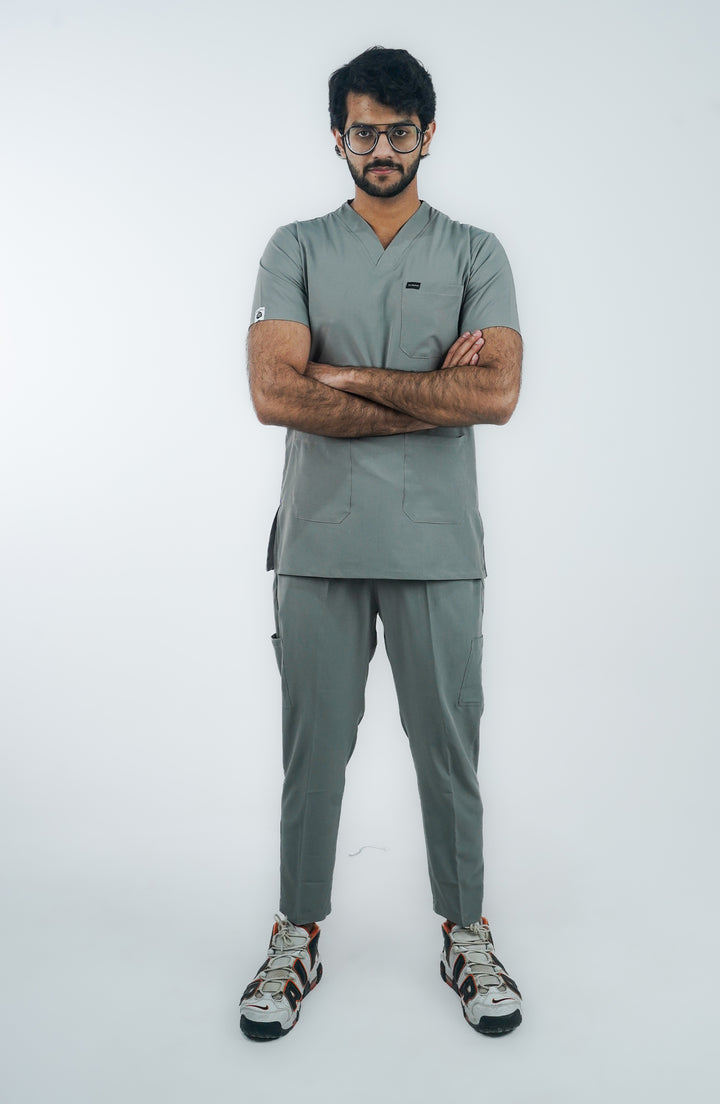 A person standing against a white background, wearing a gray micro-stretch scrub suit with short sleeves and a V-neckline, paired with white sneakers.